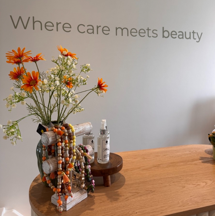 Flower arrangement with orange daisies and white baby's breath on a wooden table, beads draped over a stand, in a spa-like setting with the phrase 'Where care meets beauty' on the wall