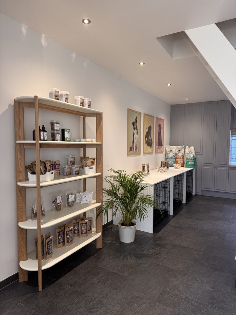 Pet store interior with a tall wooden shelf holding bags and jars on the left, a white counter and pet bags along the back wall, and framed dog pictures above the counter.